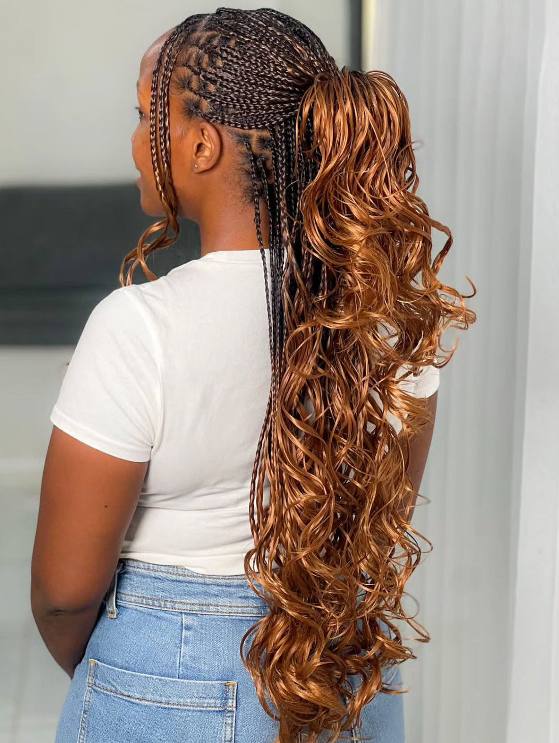 Person with braided hair styled in a ponytail with wavy extensions, wearing a white shirt and blue jeans.